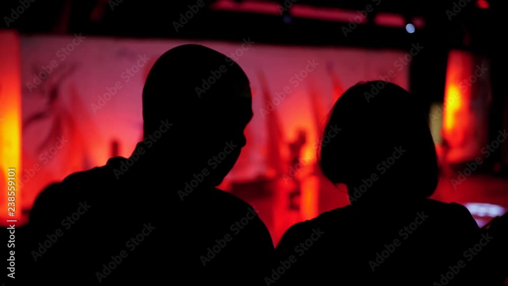 Video from the back silhouette man and woman sitting looking performance or presentation in the theater background red pink light audience concert hall stage play abstract audience slow motion