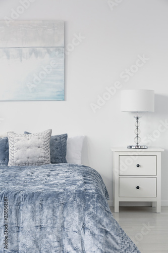 Vertical view of white stylish lamp on bedside table in luxury bedroom interior with velvet blue bedding and painting on the wall