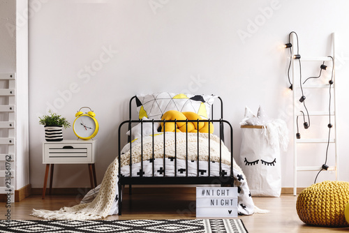 Yellow knit pillow on single metal bed with patterned duvet and white warm blanket in spacious bedroom interior with copy space on empty white wall photo