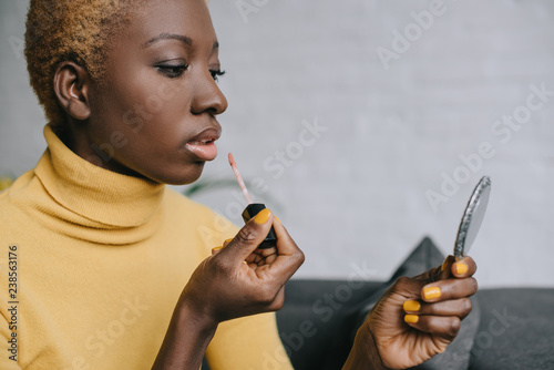 pensive african american woman applying lip gloss and looking in mirror
