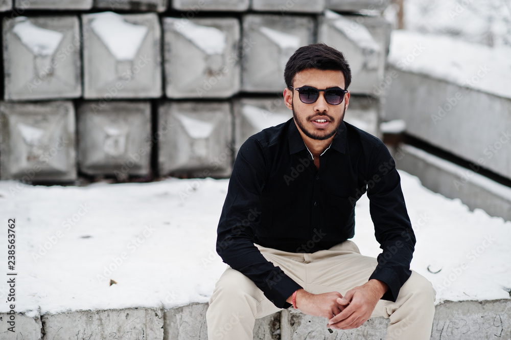 Obraz premium Casual young indian man in black shirt and sunglasses posed against stone blocks.