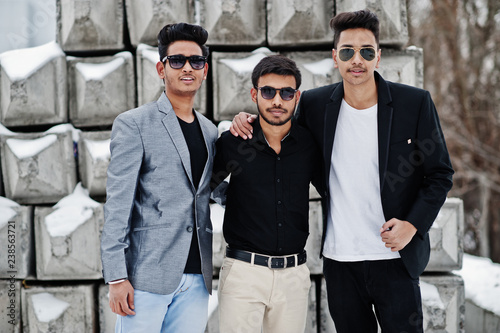 Group of three casual young indian mans in sunglasses posed against stone blocks.