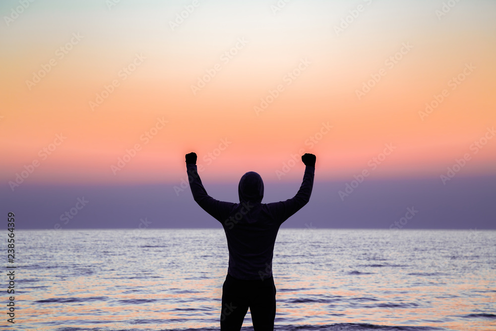 Young, strong, confidence man celebrating success after running on beach in evening. Back view. Copy space. Empty place for positive text, quote or sayings on beautiful colorful sky background.