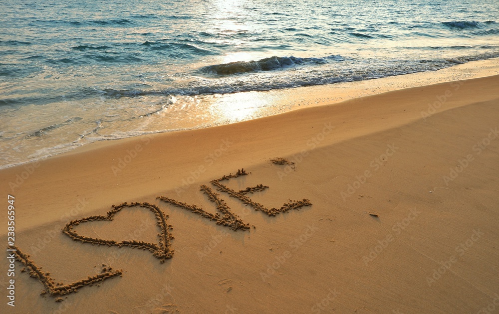 Fototapeta premium Valentines day concept, love heart shape handwriting on sand beach in sunrise and blue ocean.