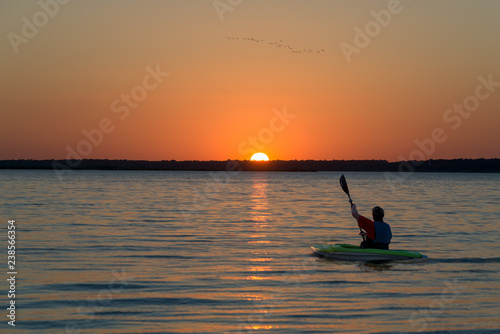Kayak rowing into a sunset at dusk on a calm lake with orange sky.