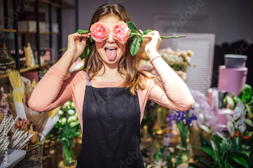 Funny and positive young female florist cover eyes with roses and show tongue. She has fun. Plants and flowers stand in vases behind.