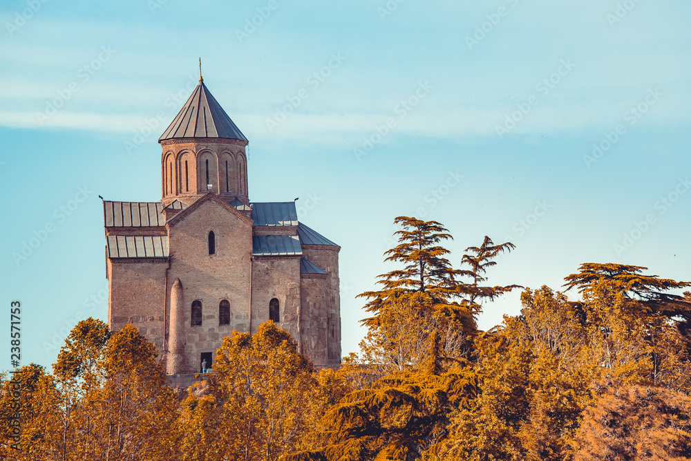 Ancient church among the autumn forest. Georgia landscape. Amazing scene of vintage Orthodox building among untouched wild nature. Blue sky background. Calm and serenity.