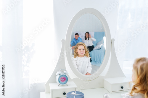 Baby girl with long hair looking at her reflection in a white room with mirror. Mother and father is in reflection