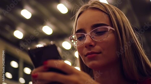 Happy young woman with glasses using cellphone at night light smile evening mobile touch phone female drink text reflection internet sms online customer smart texting slow motion