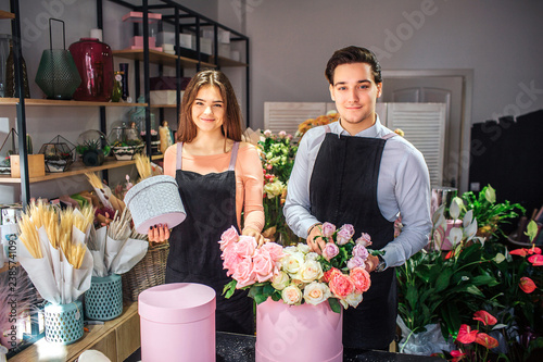 Nice and beautiful young florists look on camera. They stand at table Young woman hold small grey box. Guy touch flowers in pink box.