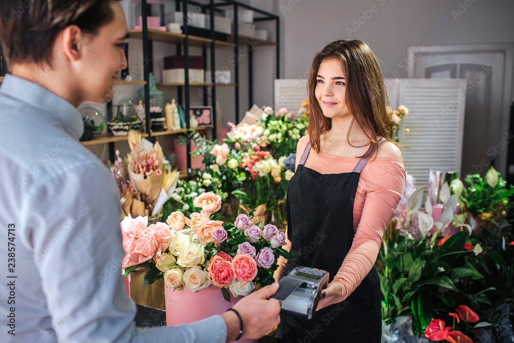 Picture of young female florist holding money therminal. She stand in front of customer. Young businessman pay for flowers.