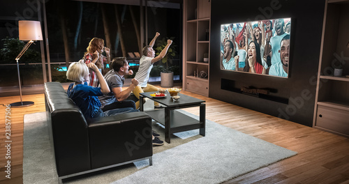 Group of students are watching a soccer moment on the TV and celebrating a goal, sitting on the couch in the living room.