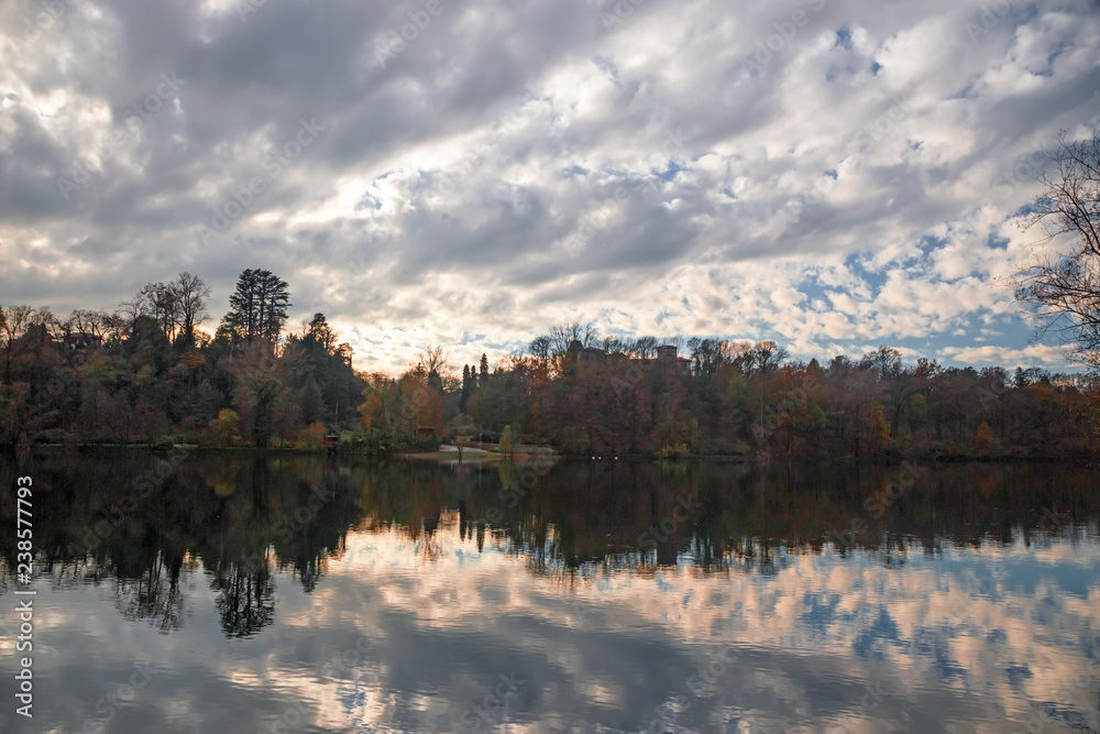 Panoramic view of the Ticino river with clouds and trees that are reflected in its clear water, on a winter day at sunset.
