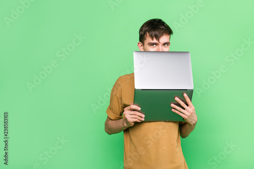 Stylish guy in a t-shirt holding a laptop Peeps out from behind the screen standing on a green background