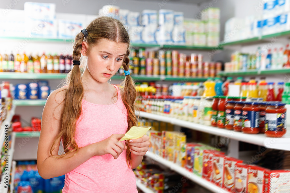 Positive tween girl choosing food products on shopping list in supermarket