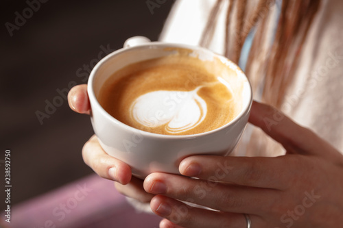 Close-up of a white Cup of hot latte art coffee with a heart shape in the hands of a young girl. Woman wearing warm winter knitted white sweater. Toned. Coffee Girl Concept Relaxation Coffee Mug.