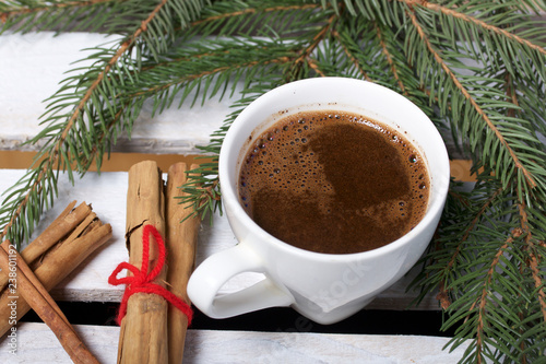 A cup of coffee and Cinnamon sticks next to the fir branches. On a wooden box.
