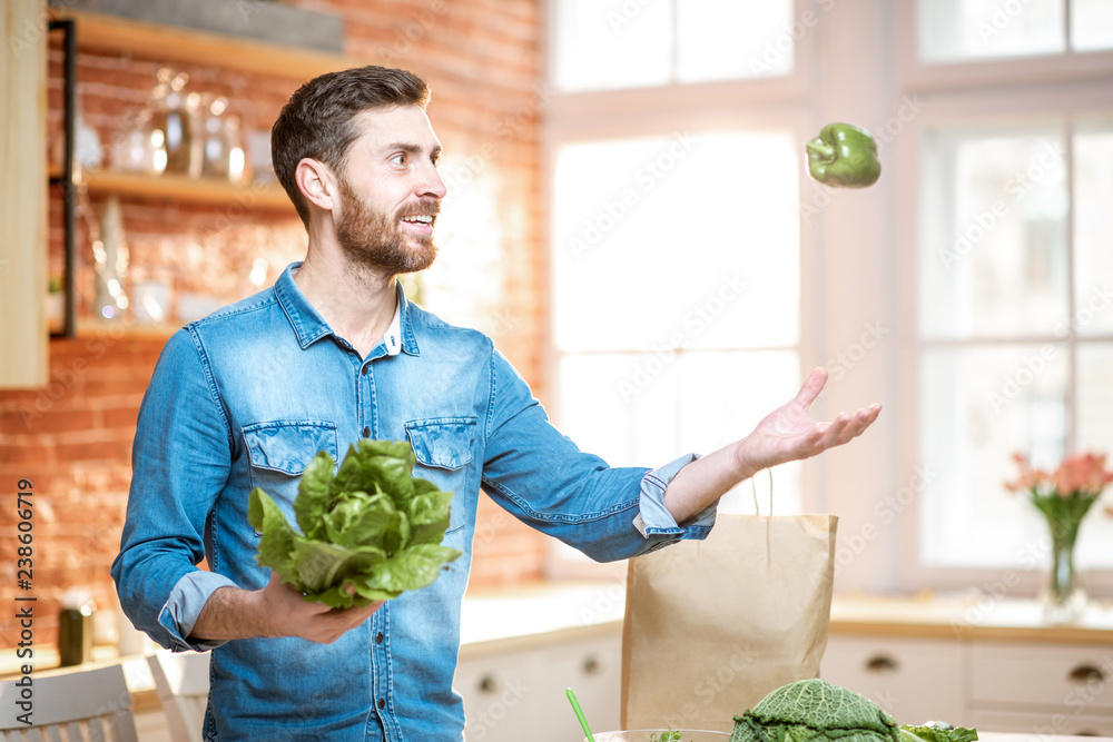 Fototapeta premium Handsome man throwing up green pepper while cooking vegan meals on the kitchen at home