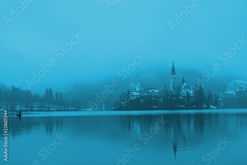 Little Island with Catholic Church in Bled Lake. With reflection of church in the lake. In blue colors. In winter in the foggy, rainy and cloudy morning. Slovenia, Bled, February 2018. With boat on th © Teya KP