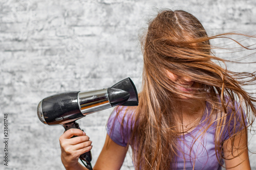portrait of young teenager brunette girl with long hair dries hair with electrical hair dryer on gray wall background