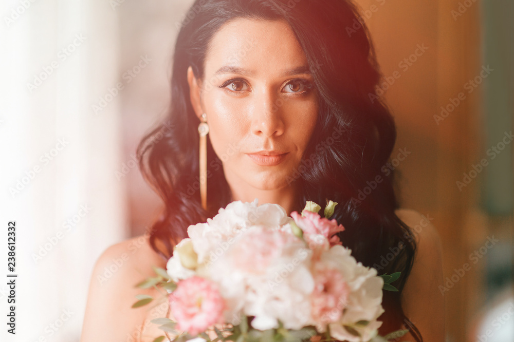 Pretty young caucasian bride with bouquet of flowers posing indoor. Lovely girl with spanish appearance closeup toned  home portrait. Dark eyed bride in white dress with pensive face tender fellings.