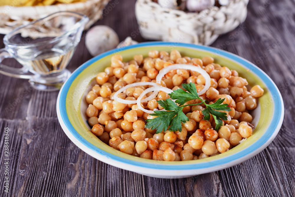 Boiled chickpeas, seasoned with vegetable oil, ground red pepper, sesame seeds and onion rings in a bowl with old spoon on a wooden background next to garlic Boiled chickpeas, seasoned with vegetable oil, ground red pepper, sesame seeds and onion rings in a bowl with old spoon on a wooden background next to garlic