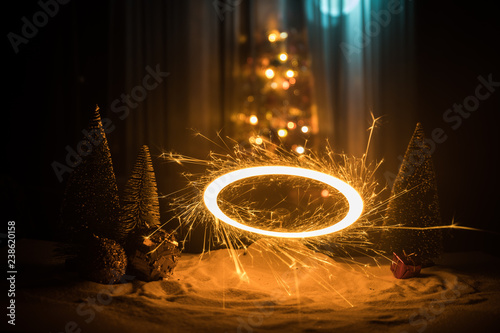 Glittering burning sparkler on snow with blurred Christmas tree on dark background. New Year Holiday concept with empty space for your text