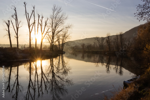 Calm and wonderful view of the river in autumn with clear reflection on the water surface, several high trees without leaves, long alley and small boat on the shore and blue sky with one plane trail