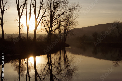 Warm sun shining through high trees without leaves on the shore of the calm river with clear reflections on the surface