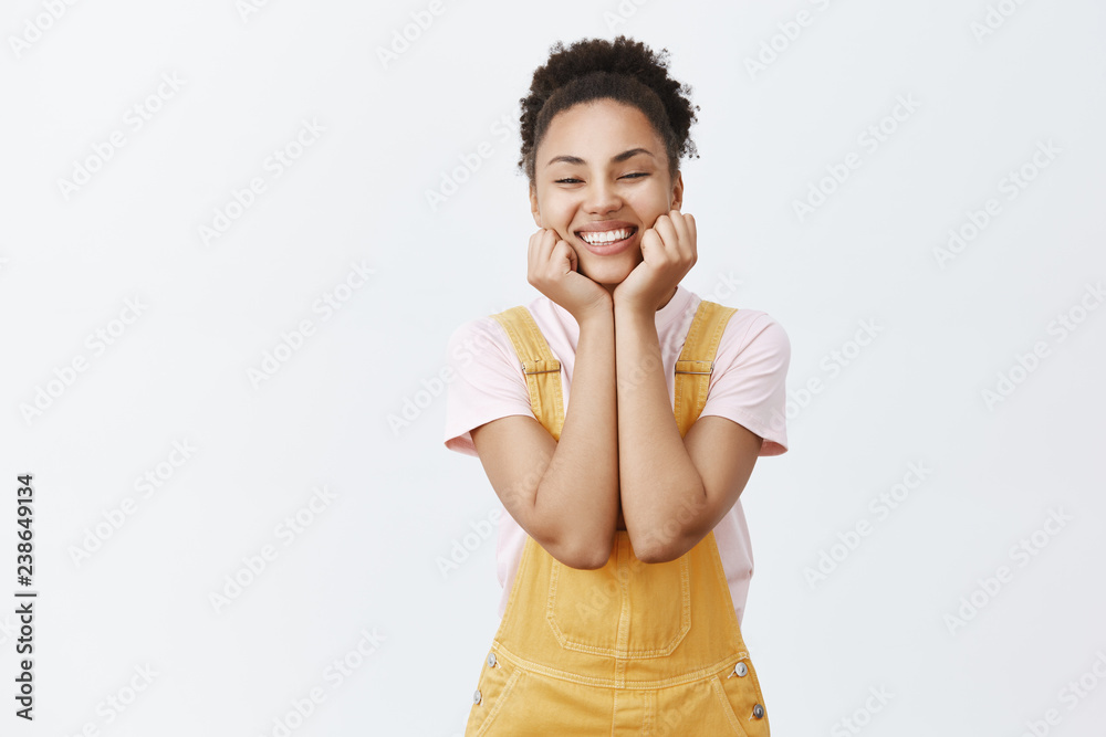 Woman being in love, telling girlfriend all details of last date. Portrait of charming african-american beautiful female in yellow trendy overalls, holding palms on chin and smiling broadly at camera Woman being in love, telling girlfriend all details of last date. Portrait of charming african-american beautiful female in yellow trendy overalls, holding palms on chin and smiling broadly at camera
