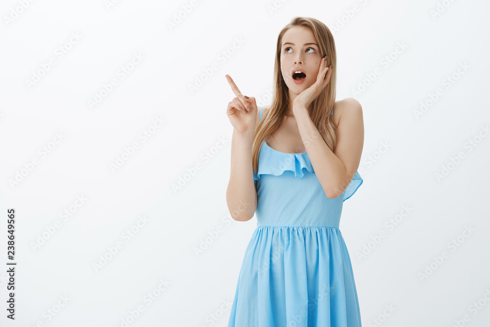 Gosh what it is. Portrait of stunned shocked and speechless attractive young blond girl in blue dress dropping jaw touching cheek and pointing at upper left corner being in stupor from amazement Gosh what it is. Portrait of stunned shocked and speechless attractive young blond girl in blue dress dropping jaw touching cheek and pointing at upper left corner being in stupor from amazement