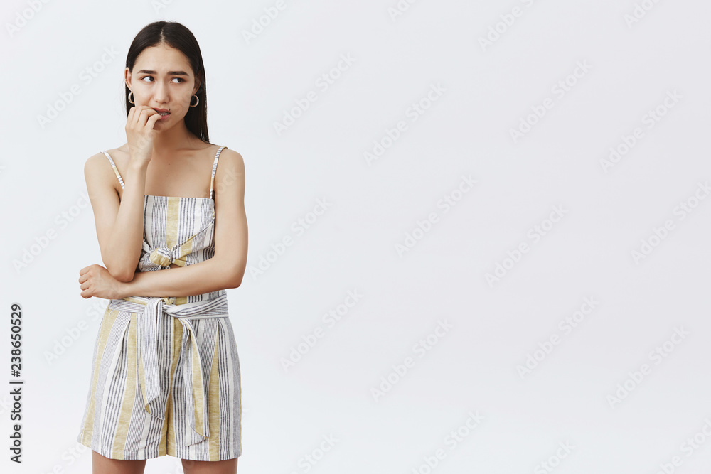 Woman always bites fingernails when nervous and distressed. Portrait of anxious attractive feminine girlfriend in overalls, biting finger and staring right with bothered worried expression, waiting Woman always bites fingernails when nervous and distressed. Portrait of anxious attractive feminine girlfriend in overalls, biting finger and staring right with bothered worried expression, waiting