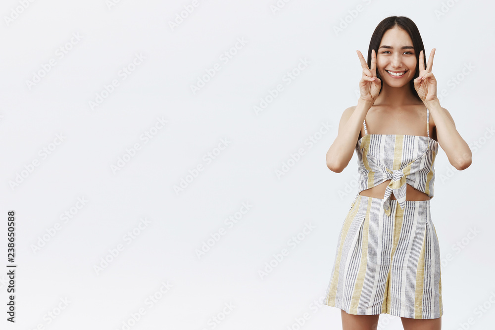 Charming friendly and stylish asian girlfriend with dark hair and cute mole under lip smiling broadly, showing peace or victory gesture near face, having fun and feeling amused over grey wall Charming friendly and stylish asian girlfriend with dark hair and cute mole under lip smiling broadly, showing peace or victory gesture near face, having fun and feeling amused over grey wall
