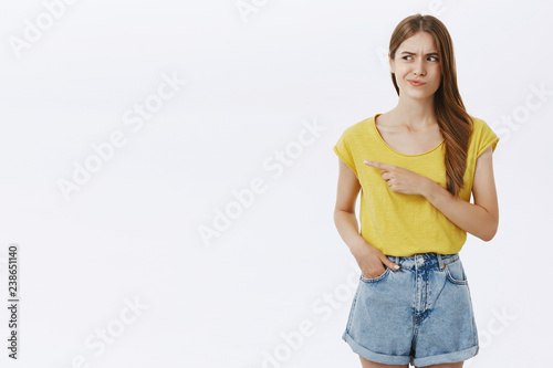 Woman mistrust suspicious seller pointing left and smirking with dislike and disbelief frowning staring at something strange holding hand in pocket of shorts posing against gray background