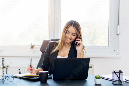 Successful young woman, smiling and taking notes while talking on the smartphone.