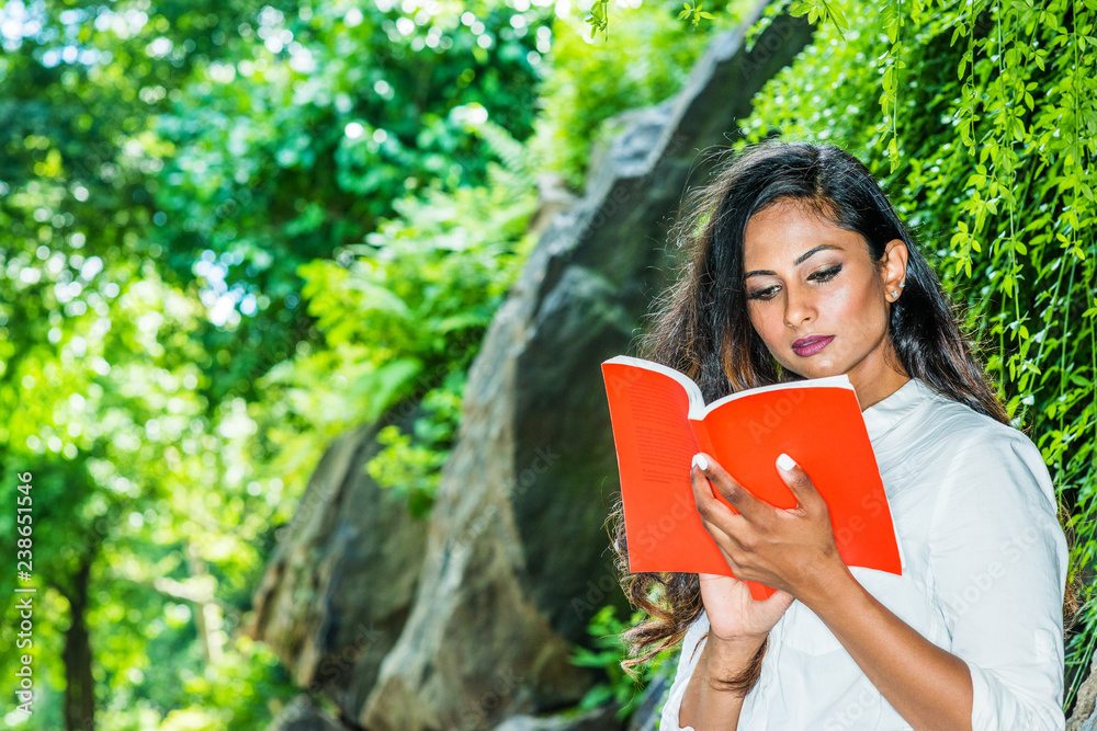 Young Beautiful East Indian American Woman with long hair relaxing outdoor at Central Park, New York City, wearing white shirt, standing by rocks with long green leaves, looking down, reading red book Young Beautiful East Indian American Woman with long hair relaxing outdoor at Central Park, New York City, wearing white shirt, standing by rocks with long green leaves, looking down, reading red book