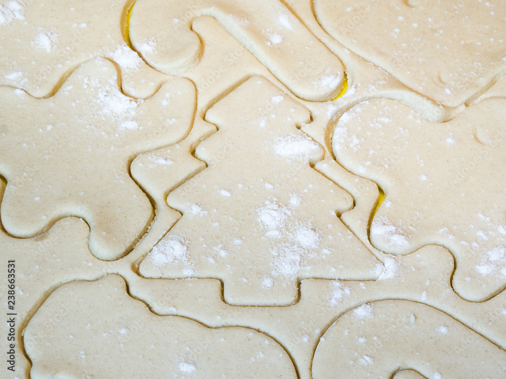A closeup of a Christmas tree cutout and other holiday stencils or shapes including gingerbread man cut in raw sugar cookie dough with flour sprinkled on top making a beautiful festive background.
