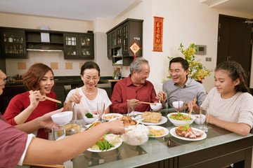 Group of cheerful Asian people enjoying good food at table served for Tet, couplets with best wishes for coming year in the background