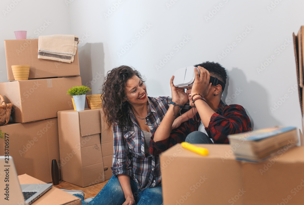 Happy couple moving into new home, they are siting on floor surrounded by packing boxes. Young man looking through virtual reality goggles.
