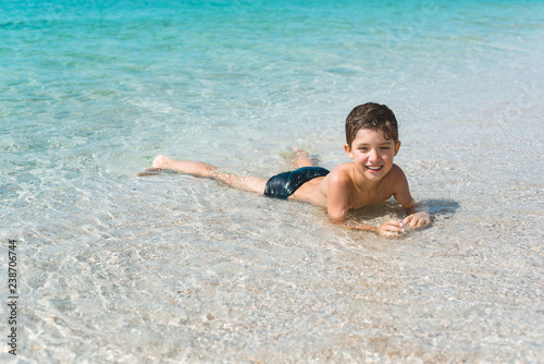A cheerful kid on the beach lies in the clear sea water.