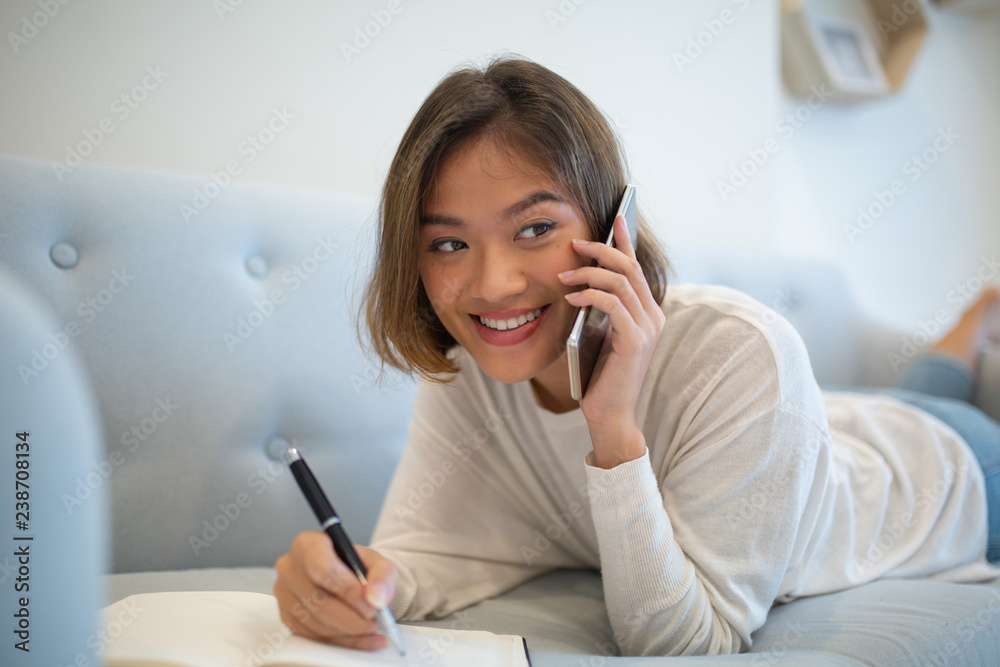 Happy lady making notes and talking on phone at home. Pretty young Asian woman studying and lying on sofa. Education and communication concept. Happy lady making notes and talking on phone at home. Pretty young Asian woman studying and lying on sofa. Education and communication concept.