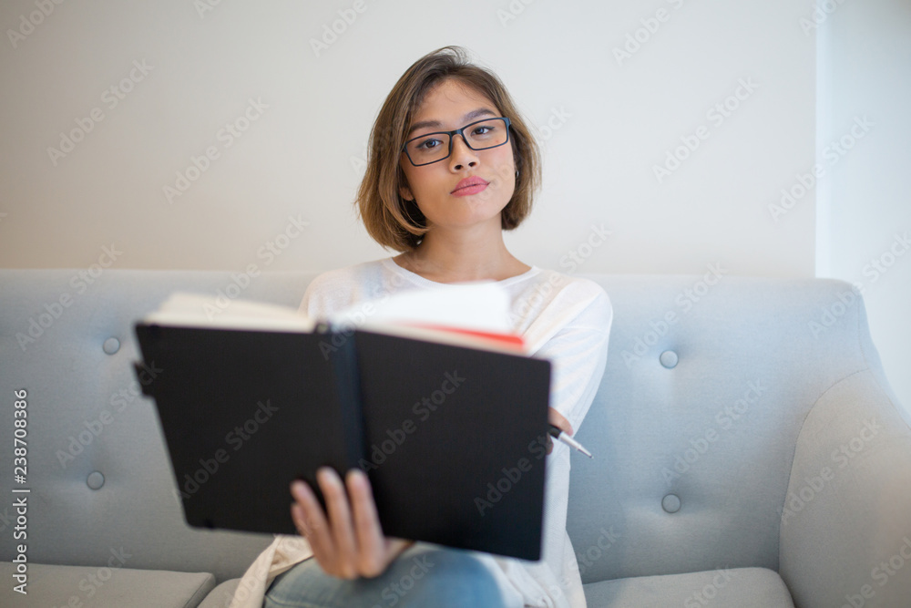 Serious Asian woman reading book on sofa at home. Pretty young lady looking at camera, sitting and holding book. Education or knowledge concept. Front view. Serious Asian woman reading book on sofa at home. Pretty young lady looking at camera, sitting and holding book. Education or knowledge concept. Front view.