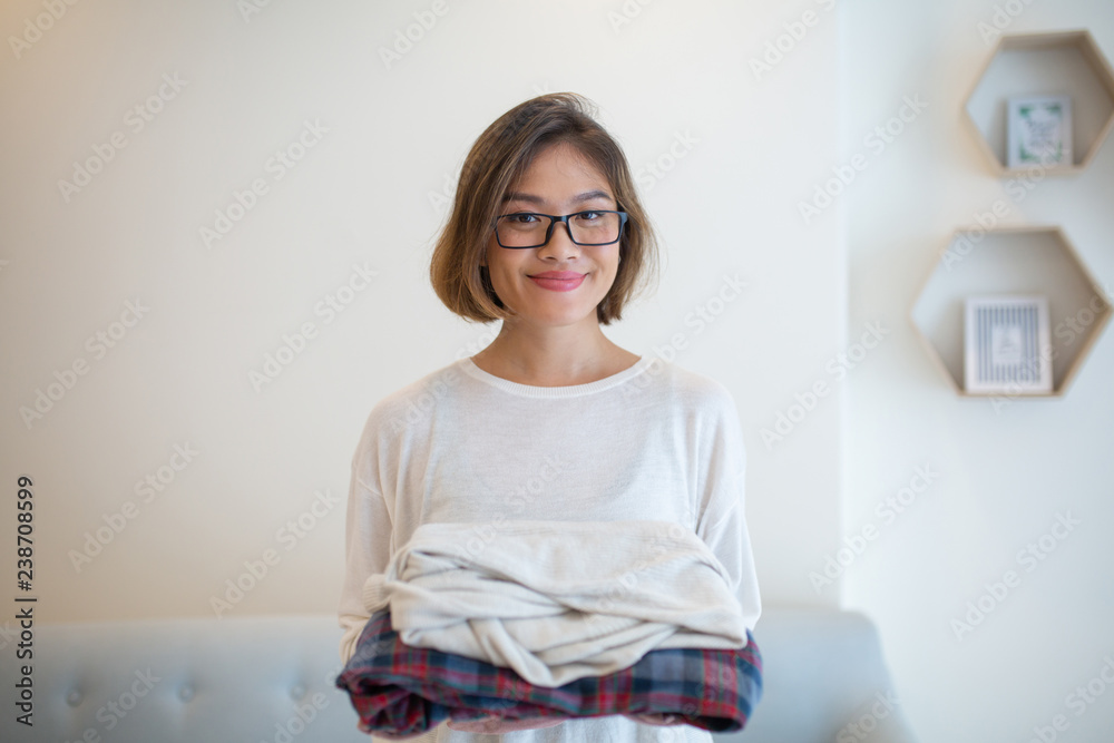 Smiling Asian woman holding clean folded clothes at home. Pretty young lady looking at camera and standing with sofa in background. Laundry and household concept. Front view. Smiling Asian woman holding clean folded clothes at home. Pretty young lady looking at camera and standing with sofa in background. Laundry and household concept. Front view.