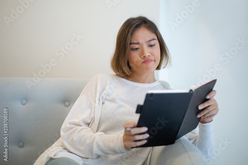 Skeptical young woman reading book on sofa at home. Pretty Asian lady sitting and studying. Education or literature concept. Front view.