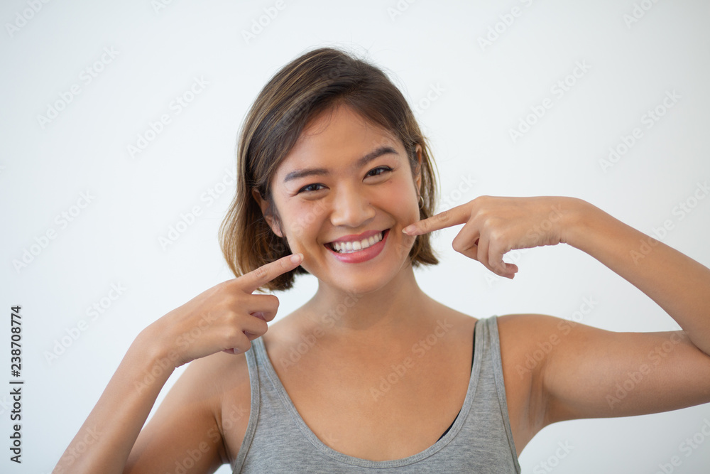 Smiling pretty Asian woman pointing at her teeth. Young woman showing teeth and looking at camera. Teeth hygiene and whitening concept. Isolated front view on white background. Smiling pretty Asian woman pointing at her teeth. Young woman showing teeth and looking at camera. Teeth hygiene and whitening concept. Isolated front view on white background.