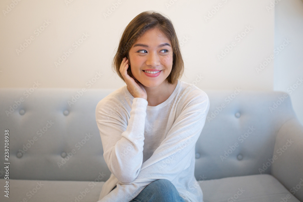 Smiling pretty young woman relaxing and sitting on sofa at home. Asian lady looking away and touching head. Leisure concept. Front view. Smiling pretty young woman relaxing and sitting on sofa at home. Asian lady looking away and touching head. Leisure concept. Front view.