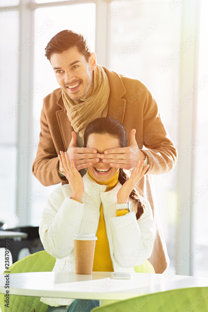 handsome man smiling and closing eyes of happy girlfriend with hands while woman sitting at table in cafe handsome man smiling and closing eyes of happy girlfriend with hands while woman sitting at table in cafe