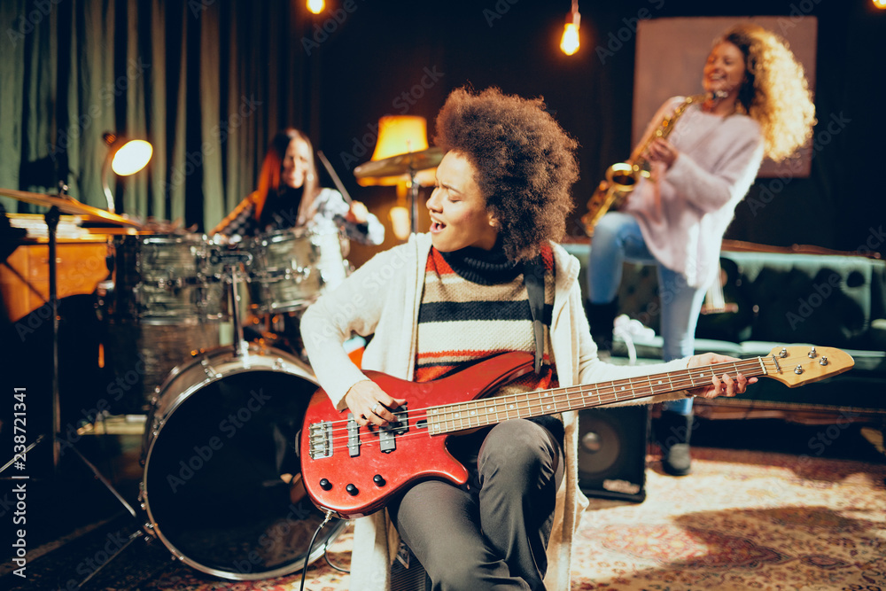 Fototapeta premium Girls playing jazz music. In foreground one woman playing bass guitar and in background other two playing saxophone and drums. Home studio interior.