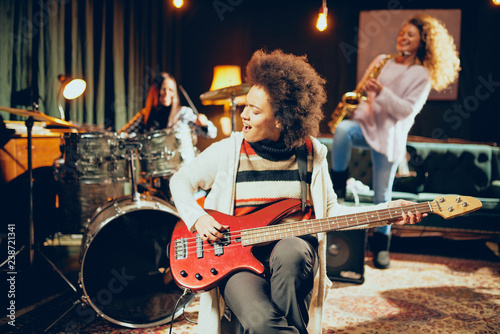 Girls playing jazz music. In foreground one woman playing bass guitar and in background other two playing saxophone and drums. Home studio interior.