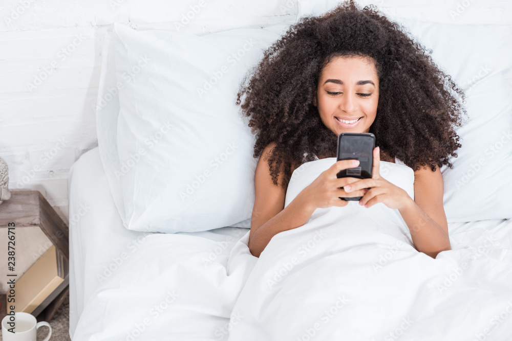 joyful african american young woman using smartphone in bed during morning time at home joyful african american young woman using smartphone in bed during morning time at home